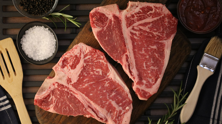 Two raw porterhouse steaks on a cutting board with BBQ sauce, pastry brush, salt & pepper, and spatula