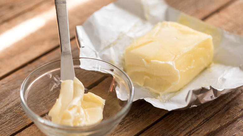 Block of butter in wrapper on wooden surface next to glass bowl with buttered knife in it