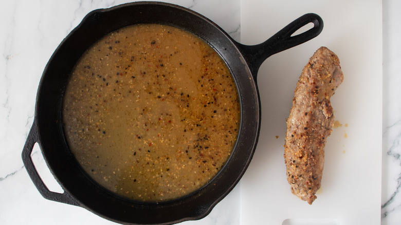 Skillet with reserved liquids next to pork tenderloin on white board