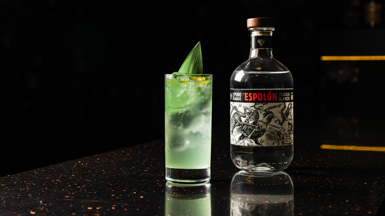 A bottle of Espolòn Blanco next to a glass of mojito on reflective countertop.
