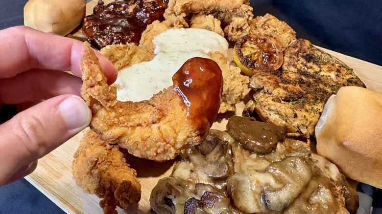 hand holding chicken tender with red sauce over wood board with more chicken