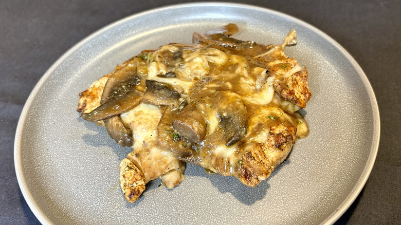 A piece of chicken covered in mushrooms, gravy, and cheese on a plate