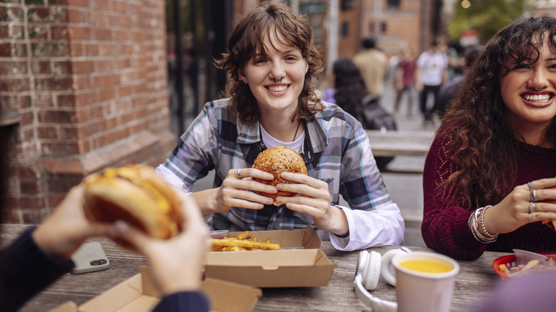 A group of friends smiling and eating burgers at a wooden table outside