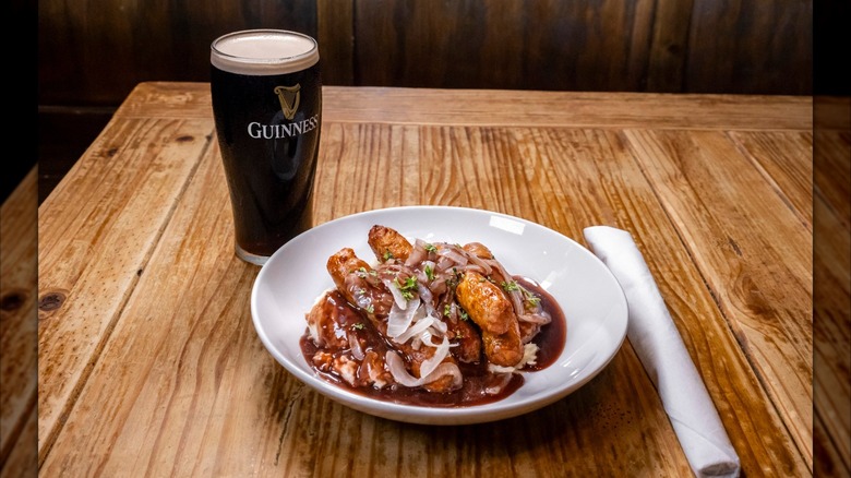 Traditional Irish dish bangers and mash on wooden table next to pint of guinness beer and roll of silverware