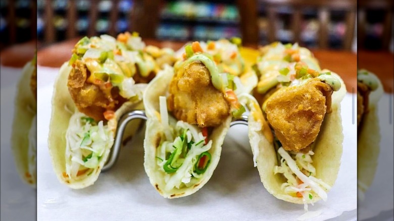 three crispy Mahi tacos in metal taco holder with restaurant blurred in background