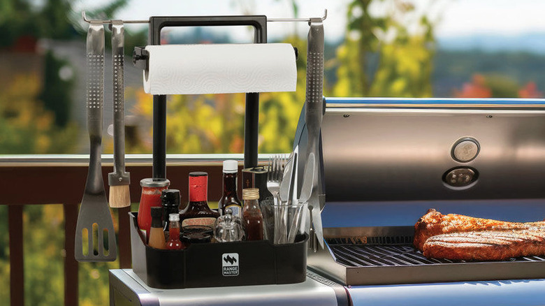 Barbecue caddy with condiments and utensils on grill next to cooking meat.