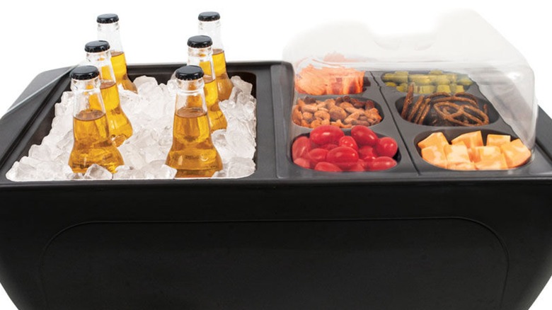 A black cooler holding six bottles of beer in ice, with snack bites including cherry tomatoes, cheese, and pretzels