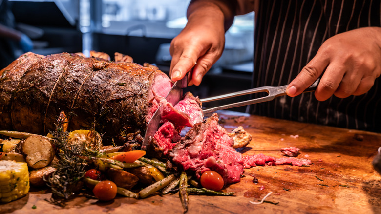 A chef slicing portions of  prime rib on a carving table alongside a bed of roasted vegetables