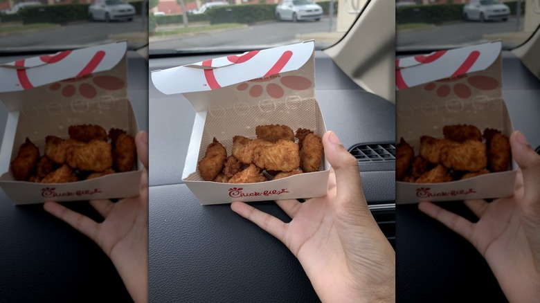 A hand holds an open takeout box of Chick-fil-A nuggets on car dashboard.