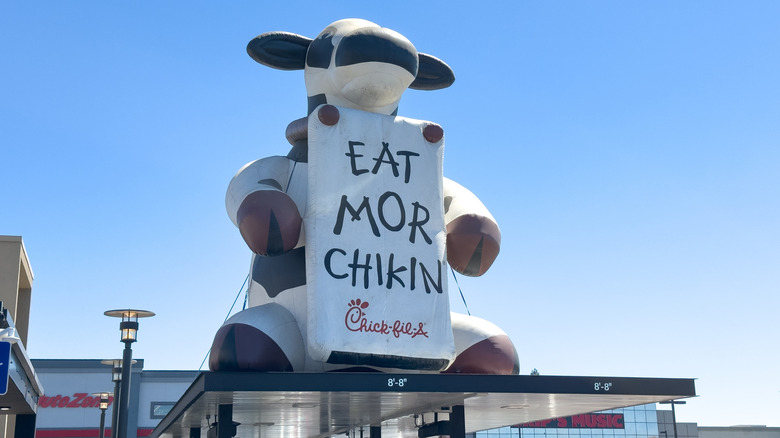 An inflatable cow sits atop a Chick-fil-A drive-thru during the day, holding a sign that reads Eat More Chikin.