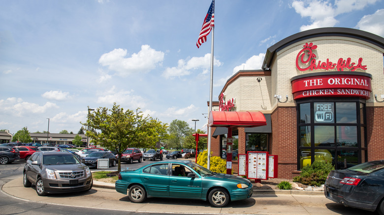 A long line of cars line wrap around Chick-fil-A restaurant in drive-thru lane during the day.