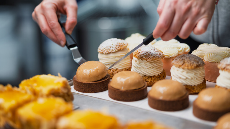 Hands arranging individual pastries on a tray, including chocolate mousse domes and cream-filled choux pastries