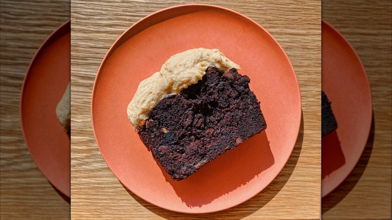 A dense slice of dark loaf cake topped with cream on the top, plated on an orange dish