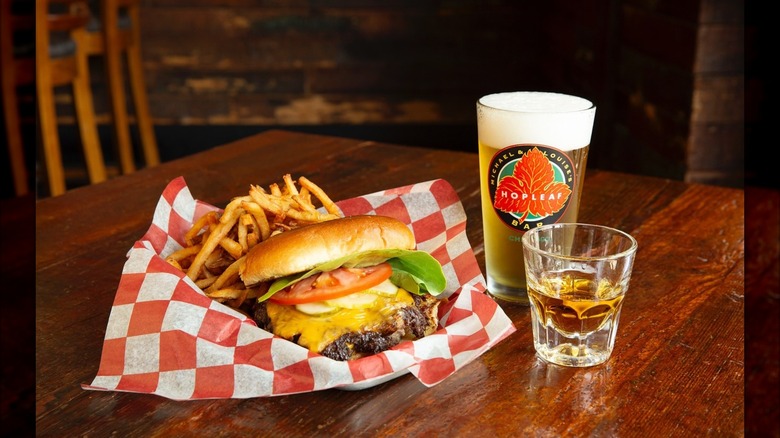 smash cheeseburger with lettuce and tomato with a side of fries in basket on wooden table next to pint of beer and shot glass of whiskey
