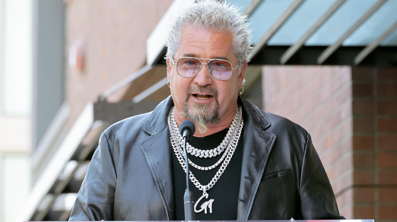 Guy Fieri in leather jacket, sunglasses, and chain necklaces stands at podium and speaks into microphone