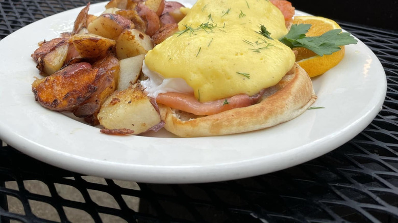 close up of eggs, English muffins, ham, home fries, and an orange slice on plate
