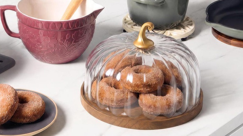 A plate, mixing bowl, pot, and pan surround a pumpkin-shaped transparent dome housing donuts.