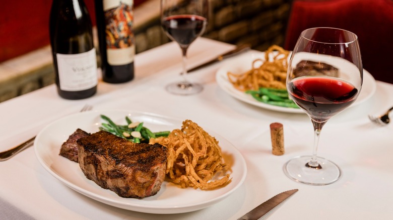 Two steak dinners on plates with fried onion strings, green beans, and red wine at a fine-dining table with tablecloth at restaurant