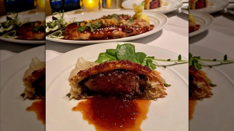 A slice of beef wellington served on a fine-dining plate with a red wine sauce and greens