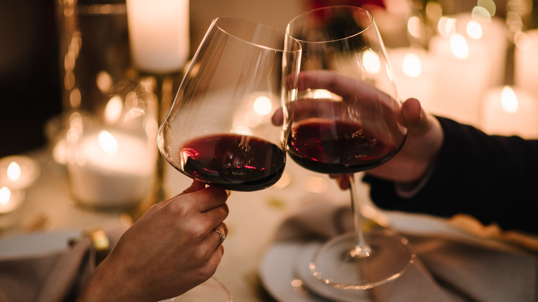 Two hands holding two glasses of red wine and clinking them together for a cheers in a dimly lit restaurant setting and candles on tables