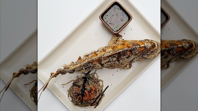 An entire fried snapper on a rectangular plate with a dipping sauce and buckwheat noodles