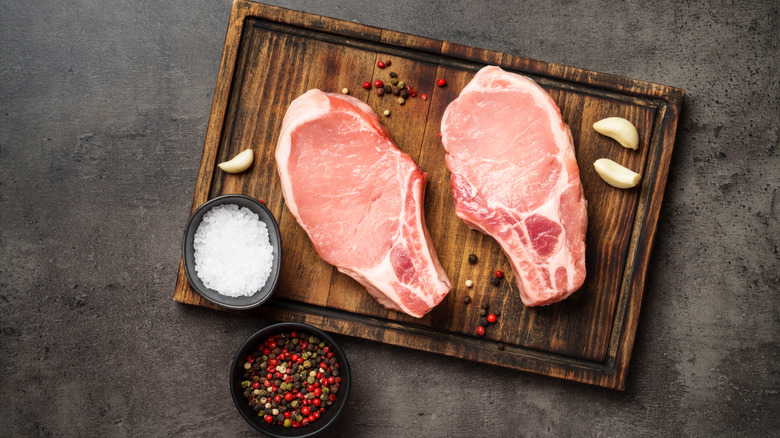 Two raw pork steaks on a wooden cutting board with herbs, salt, and garlic