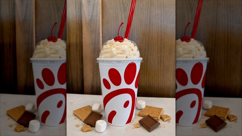 chick fil a smores milkshake topped with whipped cream and cherry in takeout cup on white counter surrounded by marshmallows, graham crackers, and chocolate pieces
