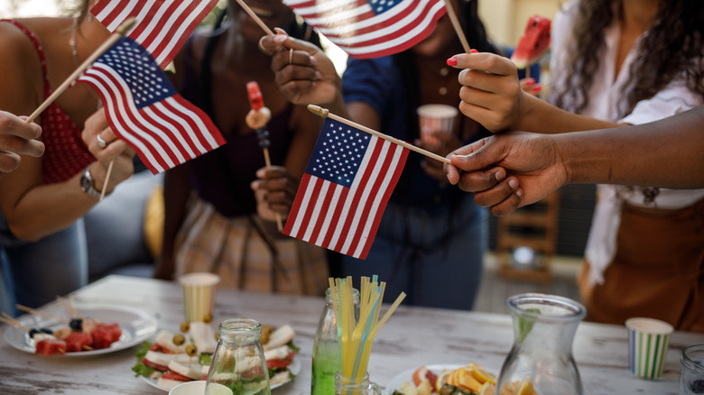People holding flags, drinks, and fruit kabobs around an outdoor picnic table