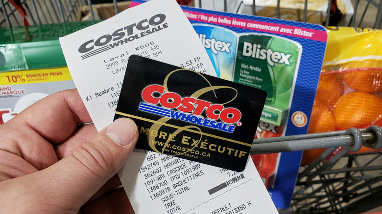 Person holding Costco membership card and store receipt over a shopping cart packed with items.
