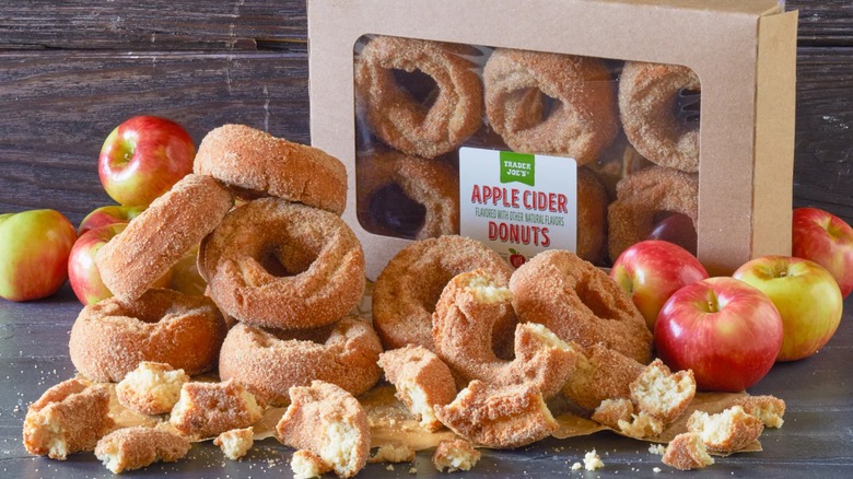 A mound of apple cider donuts on wooden table with a donut box and apples in the background.