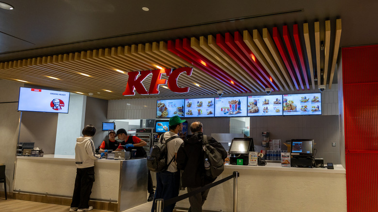 Three customers look at menu board at the counter inside a KFC
