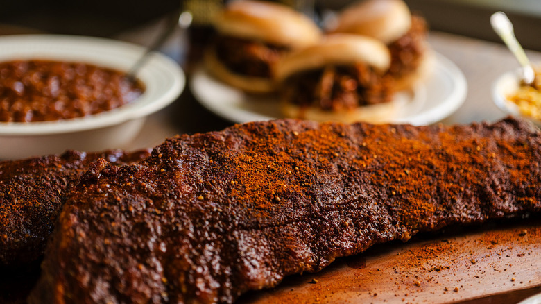 A close-up of Memphis dry rub barbecue ribs, with pulled pork sandwiches and a bowl of baked beans in the background