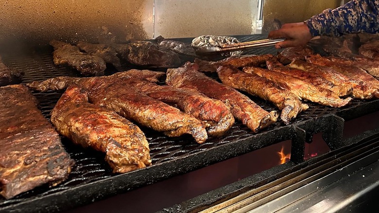 A commercial grill full of meat cooking, with a man's hand turning meat over with metal tongs