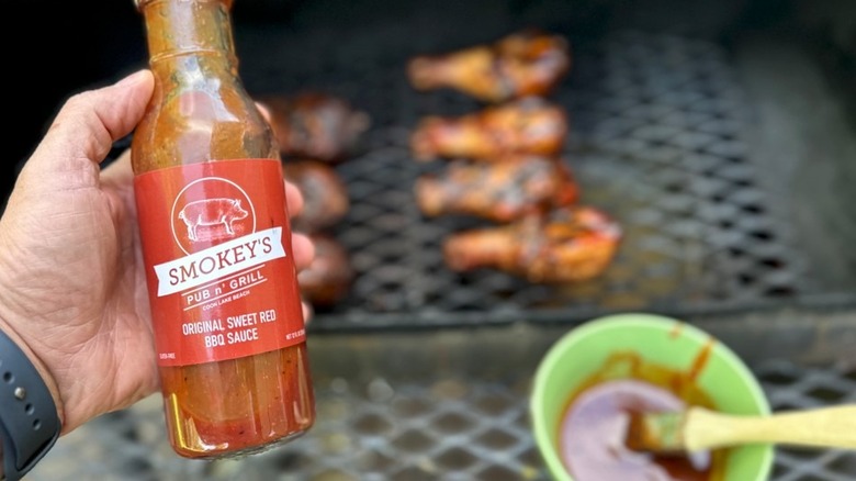 hand holding a bottle of Smokey's original sweet red BBQ sauce over a grill