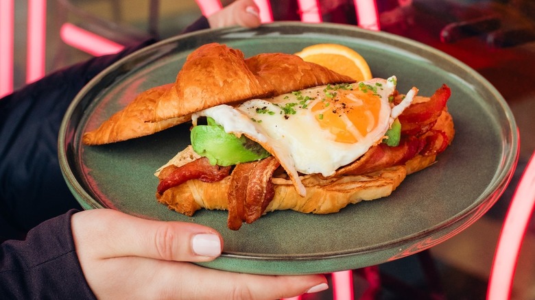 Hands holding a plate with a croissant sandwich with egg, avocado, and bacon