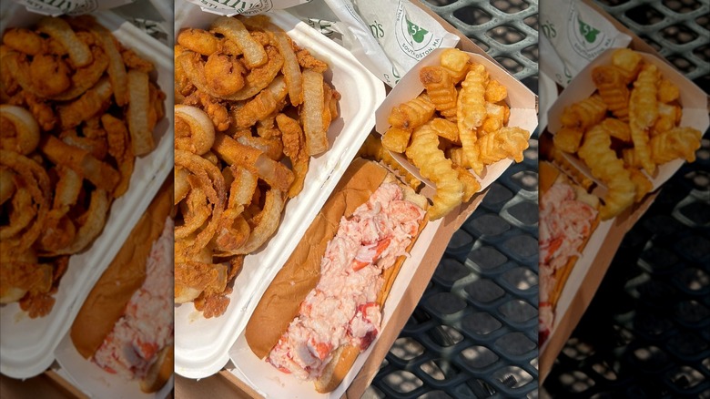 A lobster roll, onion rings, and fries in take-out containers on a table