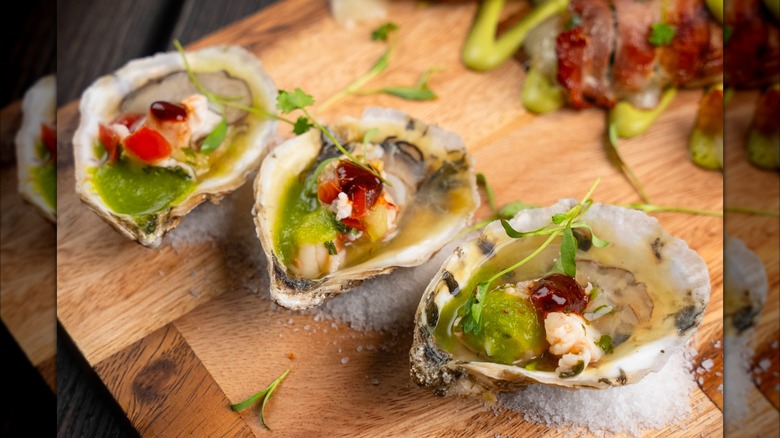 3 shucked oysters topped with shrimp ceviche, tomatillo mezcal, and sauce, on a wooden board.