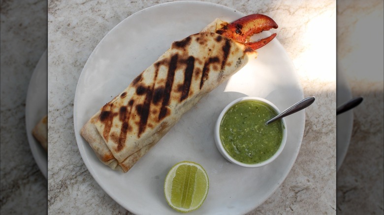 An overhead shot of a burrito with a lobster claw sticking out of one end, served with lime and green salsa