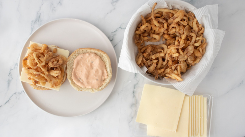 Burger buns with sauce and crispy onions next to bowl of crispy onions and cheese slices