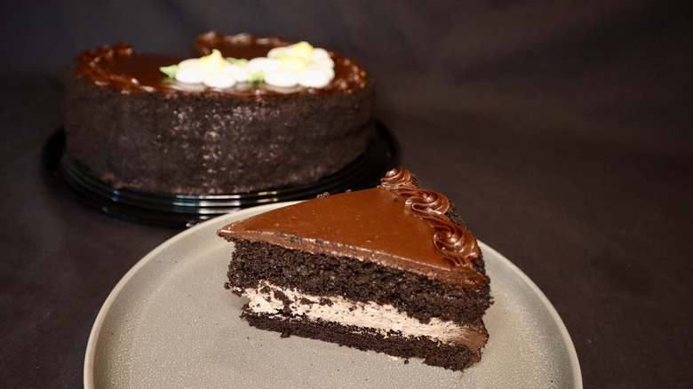 A slice of chocolate cake on a plate with a full cake in the background