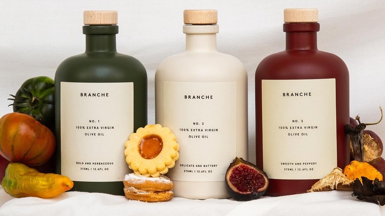 Three bottles of Branche olive oil displayed next to various cookies and vegetables