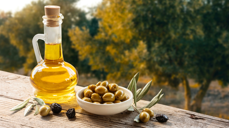 Bottle of olive oil beside bowl of olives outside with trees in the background