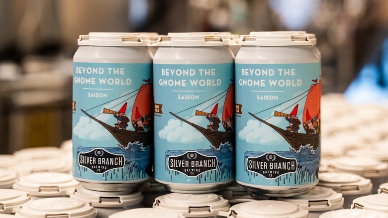Three beer cans labeled "Beyond the gnome world - saison. Silver Branch Brewing Co". The design depicts gnomes in a small boat, one using a telescope, under a blue sky