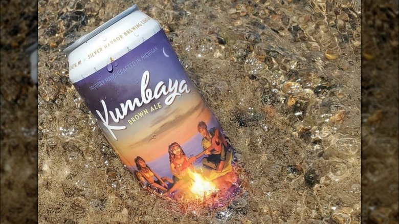 Kumbaya Brown Ale beer can partially submerged in sea water and sand at a beach. The design features people around a campfire at dusk.
