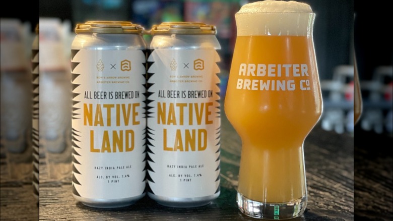 2 white cans of Native Land Beer that read "All beer is brewed on native land", alongside a pint of beer that reads "Arbeiter Brewing Co"