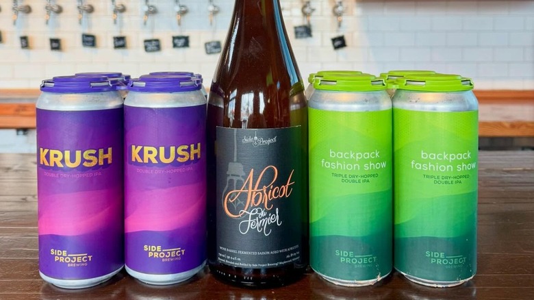 Two purple cans of Krush double dry-hopped IPA, a beer bottle with a black laber that reads "Abricot du Fermier", and 2 green cans that read "backpack fashion show triple dry-hopped double IPA".