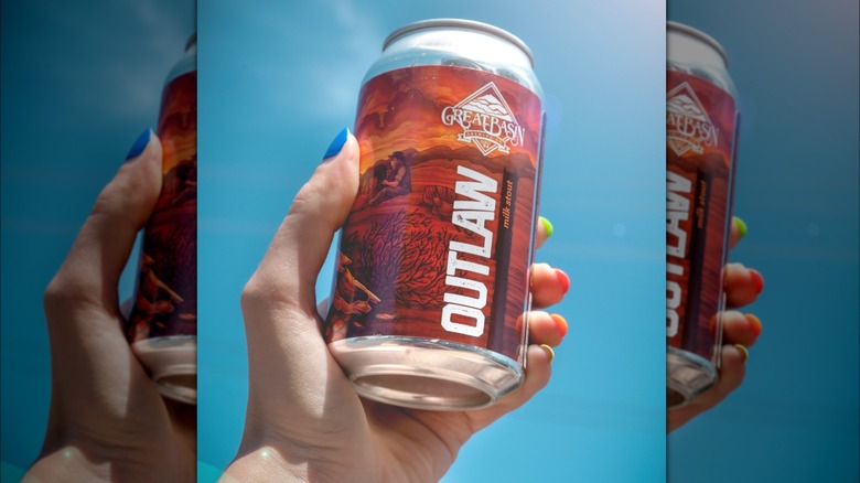A hand holding a beer can against the blue sky. The can is an Outlaw milk stout with a brown design of a man and a horse in what it looks like to be a farm, with the logo of Great Basin Brewing co on top.
