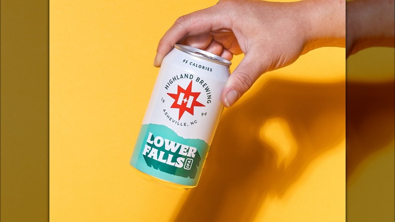 A hand holding a white and light blue beer can that reads "95 calories, Highland Brewing 1994, asheville, Lower Falls IPA, on a yellow background.