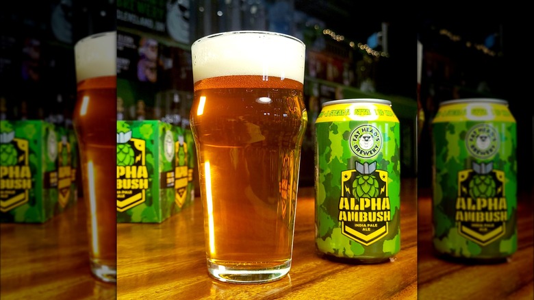 A full pint of beer alongside a beer can of Fat head brewery's Alpha Ambush India Pale Ale, with a camouflage-like design in different types of green with yellow letters.