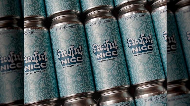 Several beer cans stacked sy by side and on top of each other. The cans read "Xul Beer Company presents: Awful nice, West Coast-style Pilsner" in a light blue design with artistic lines and flowers.